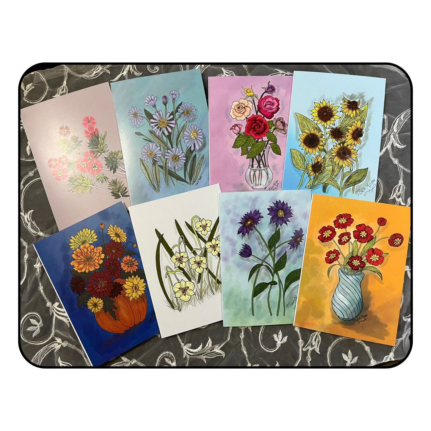 Flat lay of eight floral art note cards from Tora Moon’s Blooming Expressions set, showing all designs—sunflowers, asters, roses, fortnight lily, red cosmos, bottle brush, pumpkin bouquet, and purple daisies—arranged on a floral background.