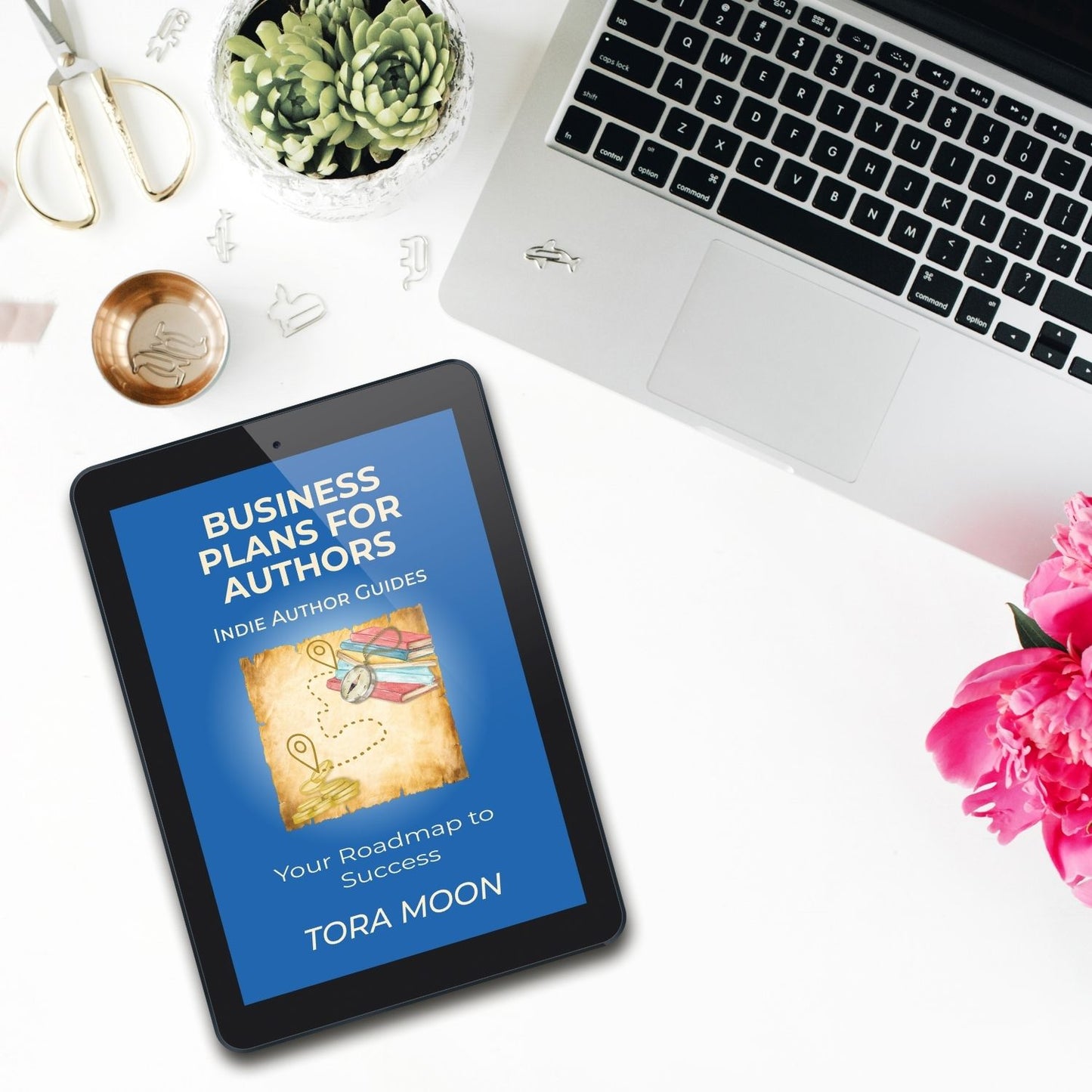 Business Plans for Authors ebook by Tora Moon displayed on a tablet beside a laptop, flowers, and writing tools. Indie author business planning guide for creative and intuitive writers.